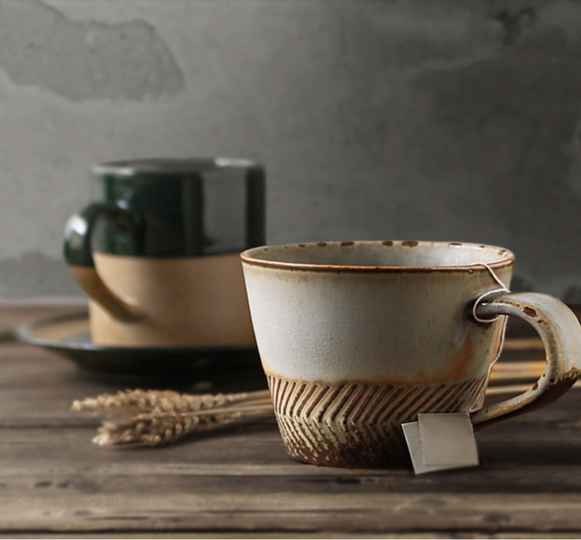 Japanese stoneware mug with carving, tan and cream, on rustic wood table with wheat stalks.